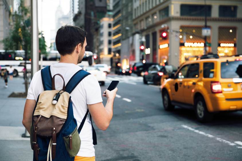 A man with a backpack stands on a city street, looking at his phone. A yellow taxi cab drives by in the background